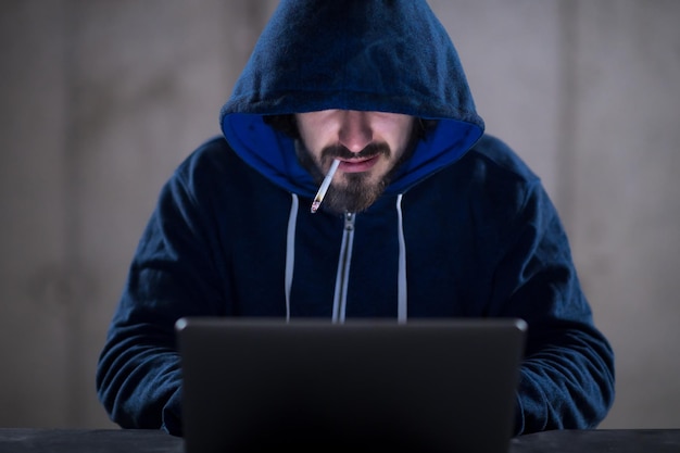 Jeune hacker talentueux fumant une cigarette tout en travaillant sur un ordinateur portable dans un bureau sombre avec un mur de béton en arrière-plan