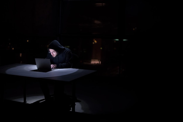 Jeune hacker talentueux utilisant un ordinateur portable tout en travaillant dans un bureau sombre avec les lumières de la grande ville en arrière-plan la nuit