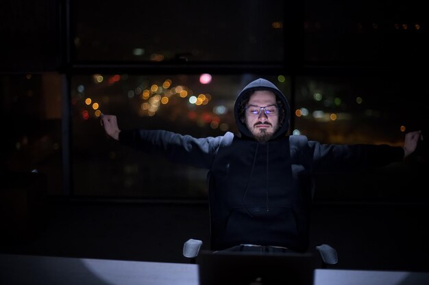 Jeune hacker talentueux utilisant un ordinateur portable tout en travaillant dans un bureau sombre avec les lumières de la grande ville en arrière-plan la nuit