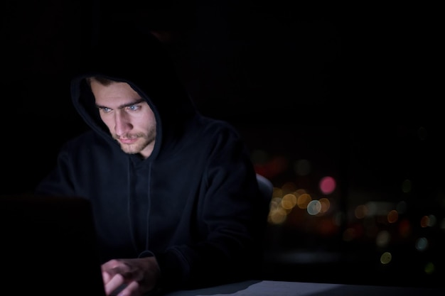 Jeune hacker talentueux utilisant un ordinateur portable tout en travaillant dans un bureau sombre avec les lumières de la grande ville en arrière-plan la nuit