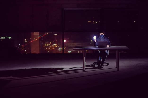 Jeune hacker talentueux utilisant un ordinateur portable tout en travaillant dans un bureau sombre avec les lumières de la grande ville en arrière-plan la nuit