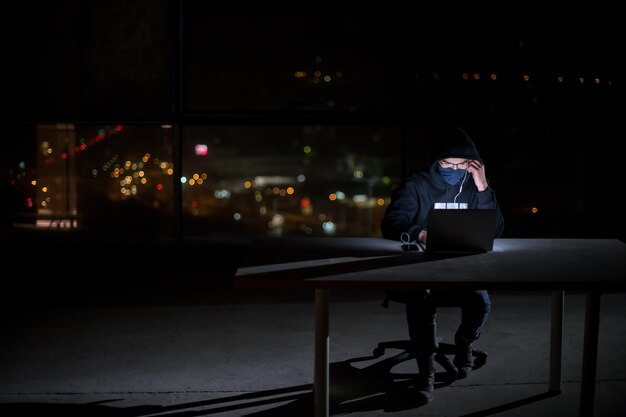 Photo jeune hacker talentueux utilisant un ordinateur portable tout en travaillant dans un bureau sombre avec les lumières de la grande ville en arrière-plan la nuit