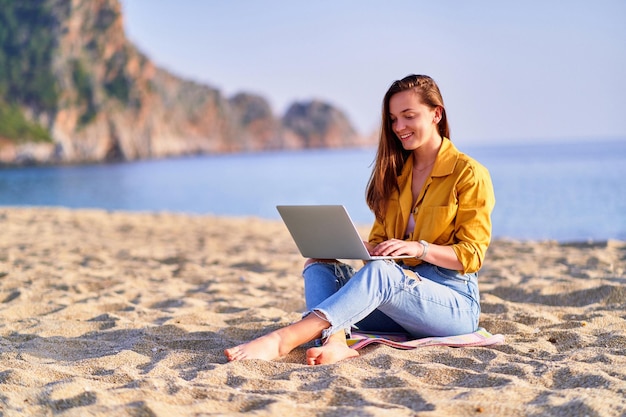 Jeune heureuse joyeuse insouciante souriante satisfaite fille indépendante du millénaire à l'aide d'un ordinateur portable sur la plage de sable au bord de la mer Concept de travail de bureau de rêve