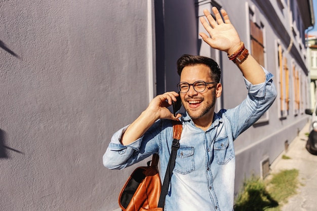 Jeune hipster attrayant debout à l'extérieur et avoir une conversation téléphonique avec sa petite amie et saluant un ami.