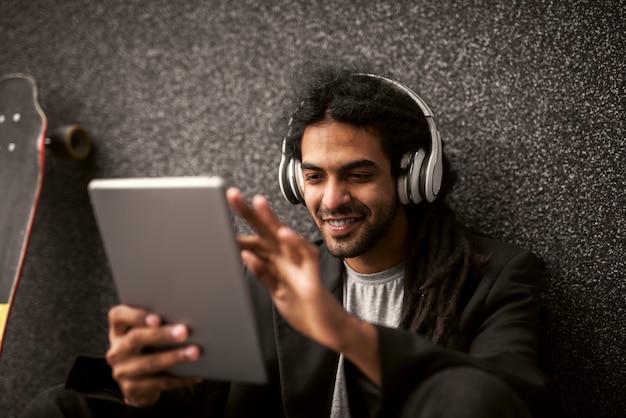 Jeune hipster dreadlock élégant avec un casque assis appuyé contre le mur gris et patinez près de lui à l'aide d'une tablette avec le sourire.