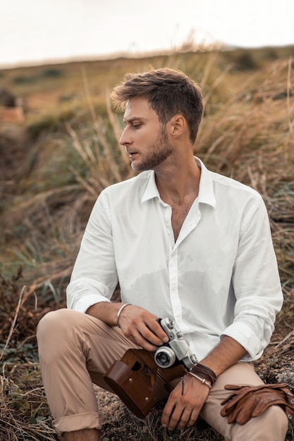 Jeune hipster mâle dans une chemise blanche avec une vieille caméra dans ses mains, posant dans la nature. Explorez des inconnus et semble cool dans des paysages étranges.