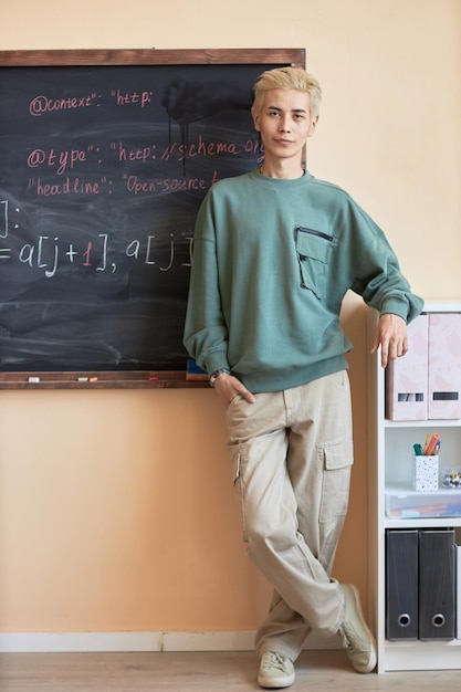 Jeune hipster masculin en pantalon gris et pull debout près du tableau noir avec des formules ou des équations écrites à la craie et regardant la caméra