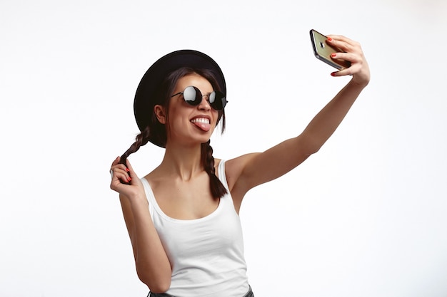Jeune hipster mignon Jeune femme prenant selfie et montrant la langue sur un mur blanc