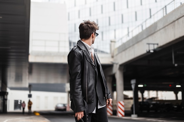 Jeune hipster à la mode dans des lunettes de soleil dans une veste en cuir noir surdimensionnée pour les jeunes avec une coiffure élégante debout et regardant en arrière près d'un bâtiment moderne à l'extérieur. Mec branché dans la rue.