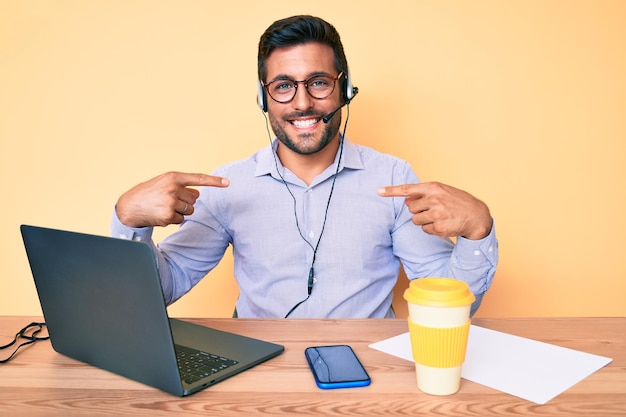 Jeune hispanique assis au bureau portant un casque d'opérateur au bureau du centre d'appels ayant l'air confiant avec le sourire sur le visage se pointant avec les doigts fiers et heureux