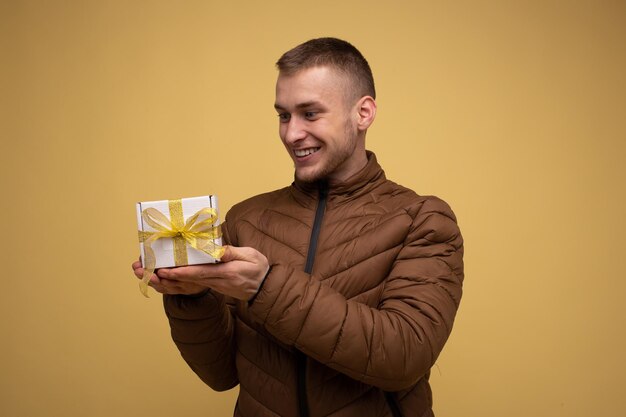 Jeune homme de 20 ans dans une veste marron sur fond jaune regardant une boîte avec un arc jaune fait d'un ruban cadeau