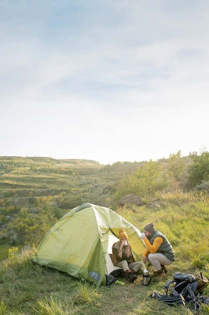 Jeune homme accroupi sur l'herbe verte par tente où sa femme se détend et prend du thé chaud après un long voyage pendant le voyage le week-end d'automne