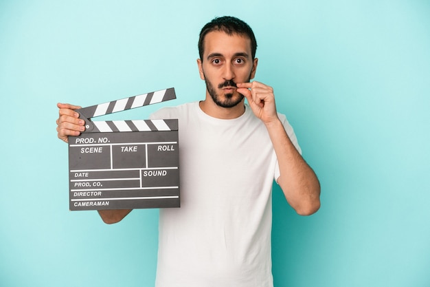 Jeune homme acteur caucasien tenant un clap isolé sur fond bleu avec les doigts sur les lèvres gardant un secret.