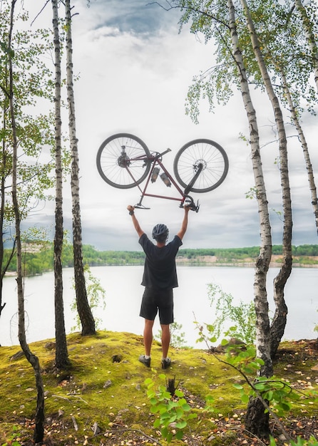 Jeune homme actif tenant un vélo au-dessus de sa tête sur le fond d'un lac dans les bois