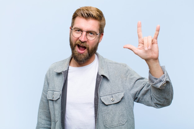 Jeune homme adulte blond se sentir heureux, amusant, confiant, positif et rebelle, faisant du rock ou du heavy metal un signe avec la main