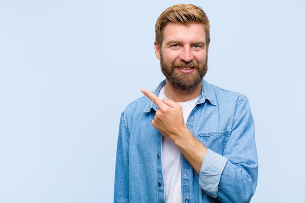 Jeune homme adulte blond souriant joyeusement, se sentant heureux et pointant vers le côté et vers le haut, montrant un objet dans l'espace de copie