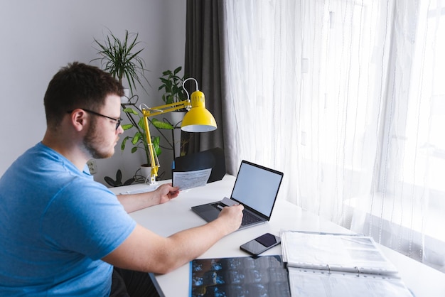 Jeune homme adulte vérifiant les résultats médicaux tenant la carte dans la main ordinateur portable avec écran blanc devant lui