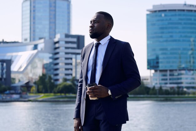 Jeune homme d'affaires africain contemporain en costume élégant prenant un verre au bord de la rivière tout en se tenant dans un environnement urbain par beau temps