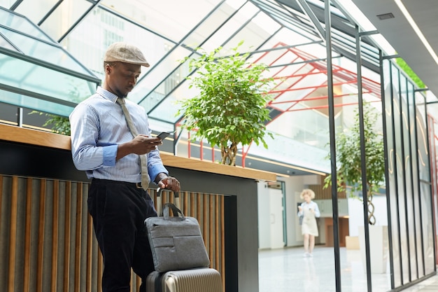 Jeune homme d'affaires africain debout avec valise et à l'aide de son téléphone portable