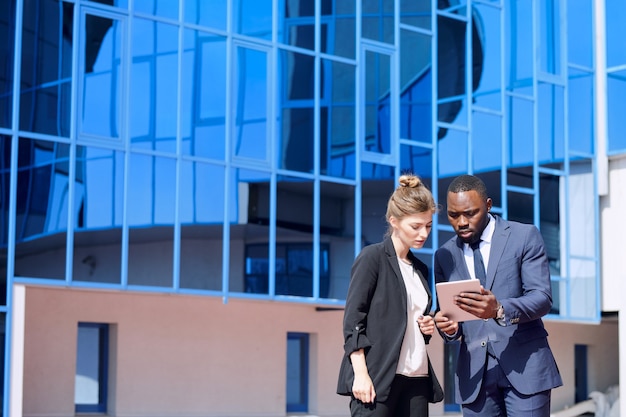 Jeune homme d'affaires africain élégant avec tablette montrant à son collègue l'horaire des vols devant la caméra contre un bâtiment moderne