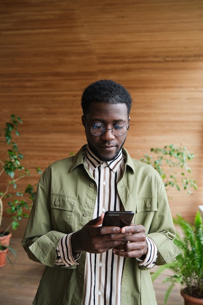 Jeune homme d'affaires africain tapant un message sur son téléphone portable en se tenant debout au bureau
