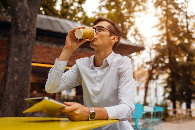 Le jeune homme d'affaires apprécie le café dans le café en plein air à l'aide d'une tablette. Beau mec buvant du thé dans la rue de la ville tenant une tablette