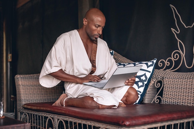 Jeune homme d'affaires arabe concentré travaillant à la pige de son costume, l'homme porte un peignoir blanc et est assis sur le divan en rotin oriental travaillant sur son ordinateur portable ; notion d'argent.