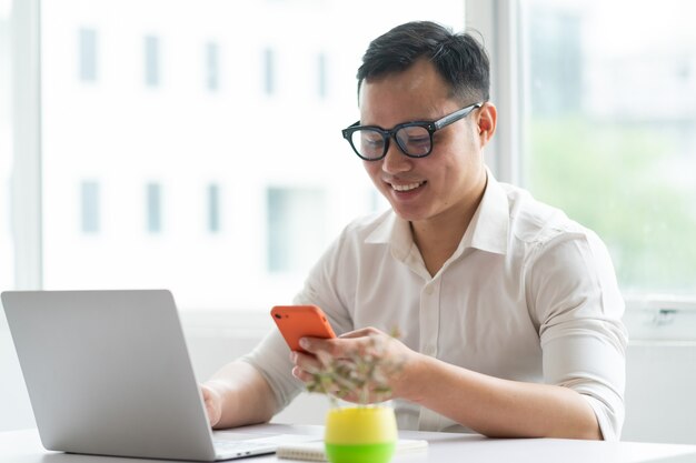 Jeune homme d'affaires asiatique à l'aide de smartphone et ordinateur portable au bureau