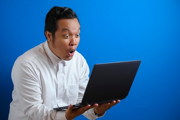 Jeune homme d'affaires asiatique portant une chemise blanche décontractée regardant un ordinateur portable, expression surprise. Bouchent le portrait du corps