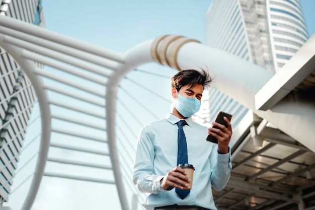 Jeune homme d'affaires asiatique portant un masque chirurgical et à l'aide d'un téléphone intelligent en ville.