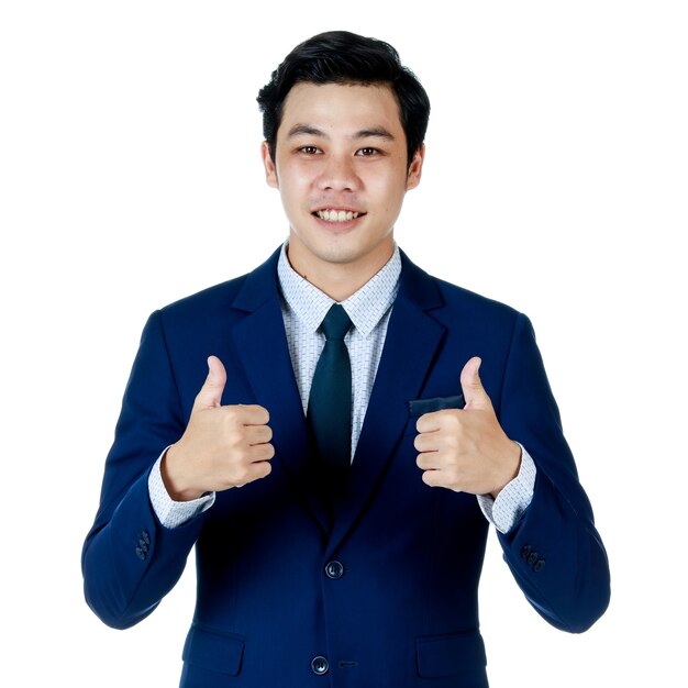 Jeune homme d'affaires asiatique séduisant portant un costume bleu marine avec une chemise blanche et une cravate, l'air confiant et levant les pouces. Fond blanc. Isolé