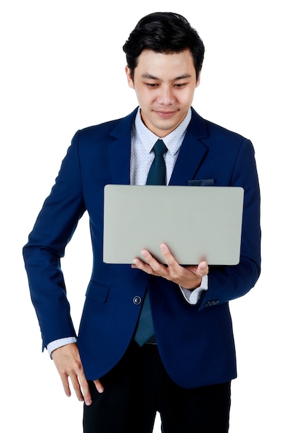 Jeune homme d'affaires asiatique séduisant portant un costume bleu marine avec une chemise blanche et une cravate tenant et utilisant son ordinateur portable avec une main dans la poche souriant sur fond blanc. Isolé