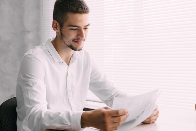 Jeune homme d'affaires assis au bureau près de la fenêtre examine des documents sur papier Le responsable du service de crise a terminé le travail et a terminé le rapport Concept de travail d'entreprise et de bureau