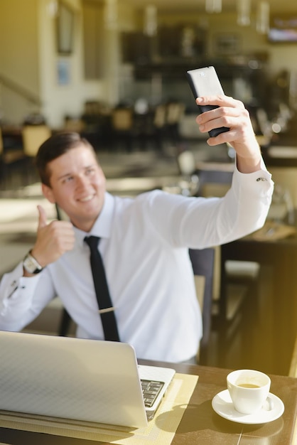 Jeune homme d'affaires assis le matin dans un café avec un ordinateur portable et une tasse de café et faisant des selfies