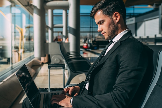 Jeune homme d'affaires assis sur l'ordinateur avec la valise à l'aéroport en attendant le vol