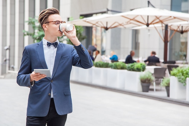 Jeune homme d'affaires attrayant avec tablette et café dans les mains sur fond de café dans la rue. tenant une tablette et buvant du café.