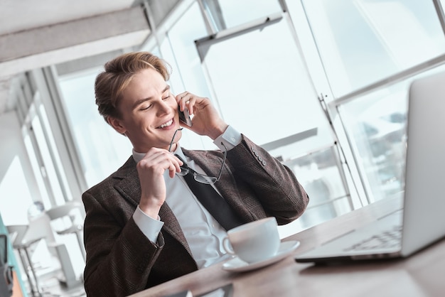 Jeune homme d'affaires au bureau assis à table avec une tasse de café chaud tenant des lunettes répondant à un appel téléphonique souriant joyeux