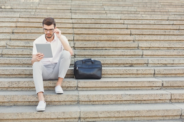 Jeune homme d'affaires ou avocat pensif lit sur une tablette numérique et touche ses lunettes tout en étant assis sur des escaliers en pierre à côté d'une mallette, copiez l'espace. Concept commercial et technologique