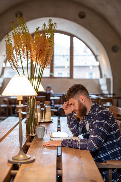 Un jeune homme d'affaires barbu est assis dans un café à table et écrit dans un ordinateur portable près de la tablette de mensonges.