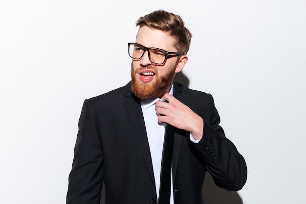 Jeune homme d'affaires barbu à lunettes et costume corrige la cravate avec la bouche ouverte et regarde de côté. Fond gris isolé