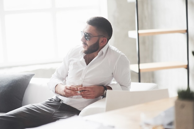 Jeune homme d'affaires barbu en lunettes de soleil et whisky à la main s'asseoir sur le canapé blanc