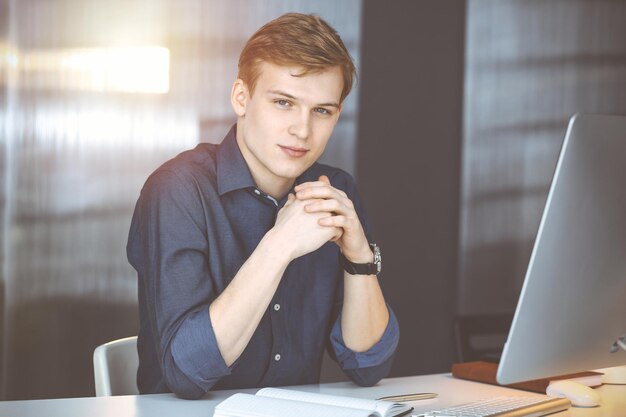 Jeune homme d'affaires blond réfléchissant à la stratégie sur son lieu de travail avec ordinateur dans un bureau sombre, éclat de lumière en arrière-plan.