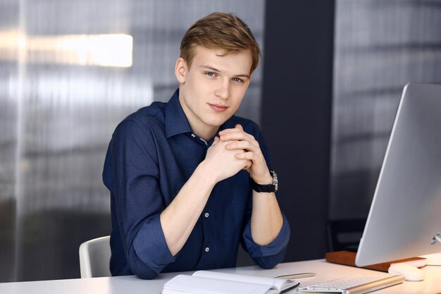 Jeune homme d'affaires blond réfléchissant à la stratégie sur son lieu de travail avec ordinateur. Une entreprise en démarrage signifie travailler dur et hors du temps pour réussir.