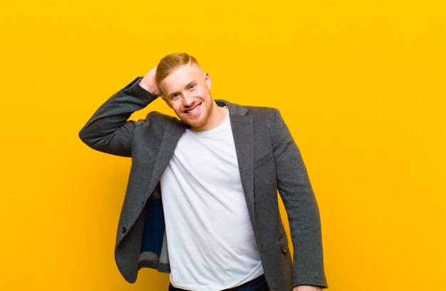 Jeune homme d'affaires blond souriant gaiement et nonchalamment, prenant la main à la tête avec un regard positif, heureux et confiant