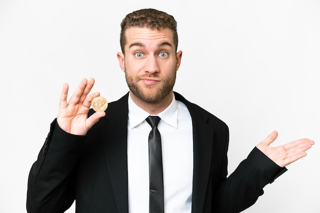 Jeune homme d'affaires blond tenant un Bitcoin sur fond blanc isolé ayant des doutes tout en levant les mains