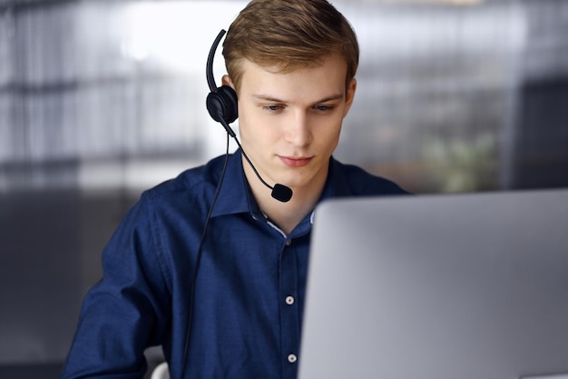 Jeune homme d'affaires blond utilisant un casque et un ordinateur au travail. Une entreprise en démarrage signifie travailler dur et hors du temps pour réussir.