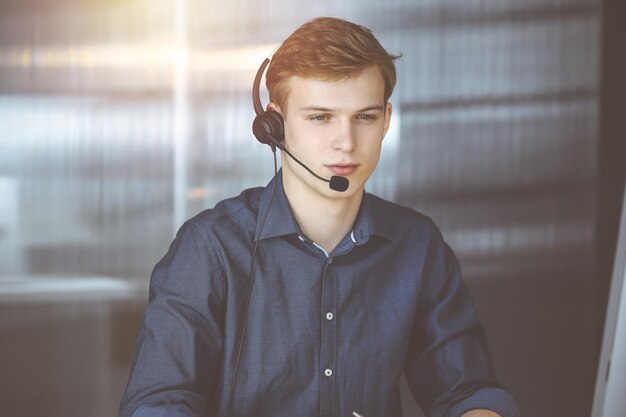 Jeune homme d'affaires blond utilisant un casque et un ordinateur dans un bureau sombre, éclat de lumière sur le fond. Démarrer une entreprise signifie travailler dur.