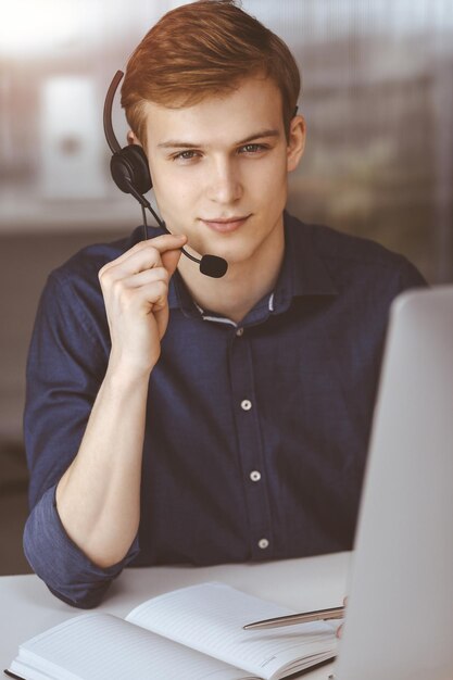 Photo jeune homme d'affaires blond utilisant un casque et un ordinateur dans un bureau sombre, reflet de la lumière sur l'arrière-plan une entreprise de démarrage signifie travailler dur