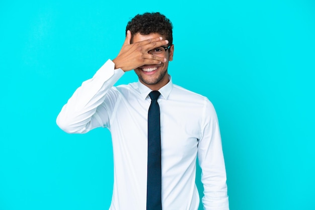 Jeune homme d'affaires brésilien isolé sur fond bleu couvrant les yeux par les mains et souriant