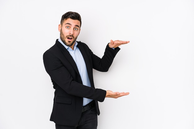 Jeune homme d'affaires caucasien contre un mur blanc isolé choqué et étonné tenant un espace de copie entre les mains.